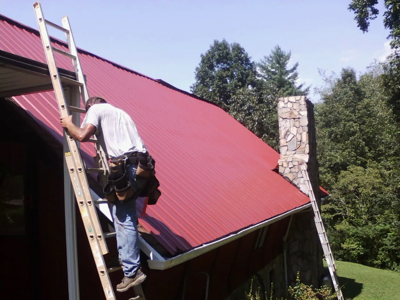 Roofer on a ladder beside a red metal roof and stone chimney — Soffit & Fascia Repair by Roofing Brothers in Oldtown